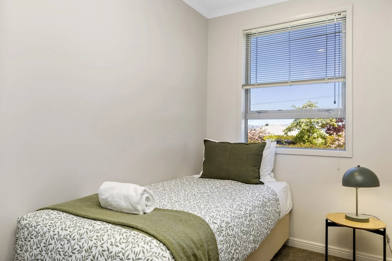 A small bedroom with a single bed covered in a leaf-patterned duvet, a rolled white towel on it, a green pillow, a hanging lamp on a small wooden table, and a window with blinds showing trees outside.