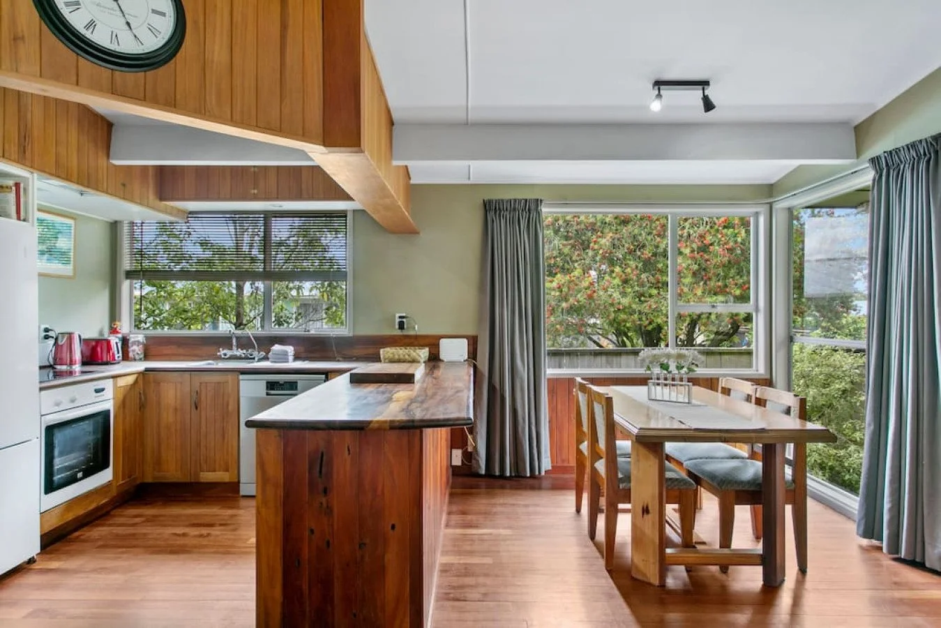 Kitchen and dining area with wooden cabinets, a window with blinds, a countertop, a stove, and a dining table with chairs near large windows showing trees outside.