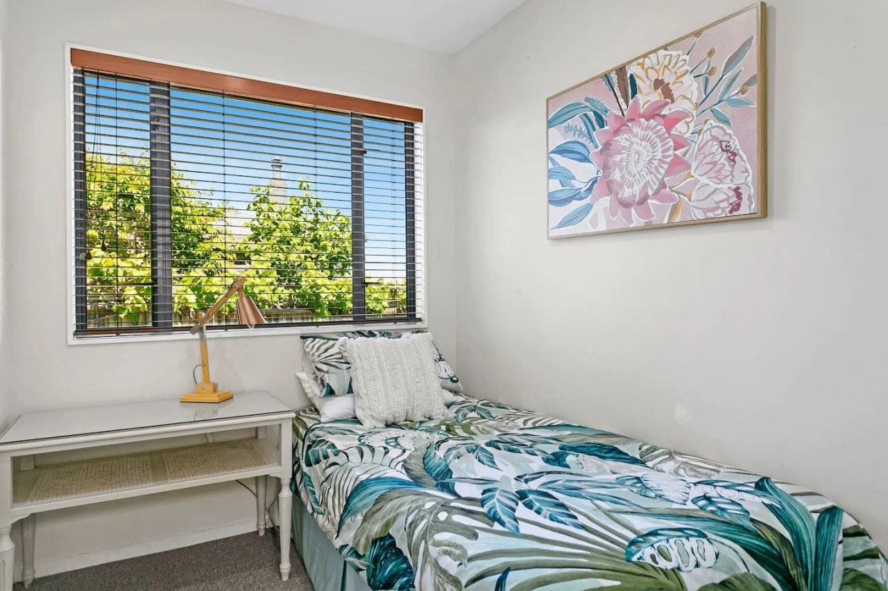 A small bedroom with a single bed that has a tropical leaf print blanket and pillows. A white side table with a yellow lamp is next to the bed. A large window with wooden blinds shows green trees outside. A floral artwork hangs on the white wall abov