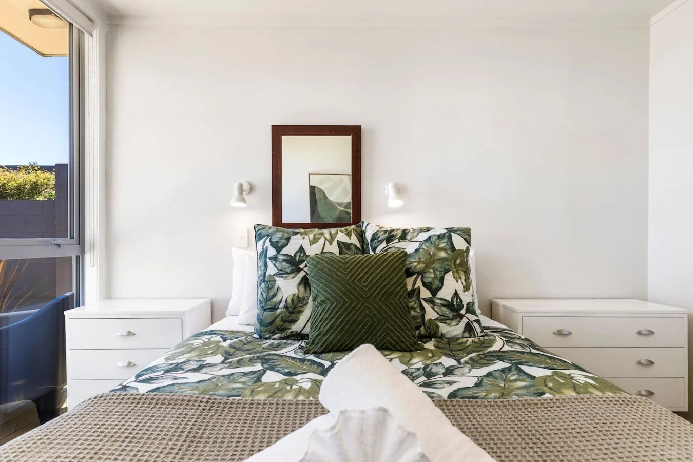 Bedroom with bed featuring botanical print bedding, green pillows, bedside tables, mirror, and white walls, with open window showing outdoor view.