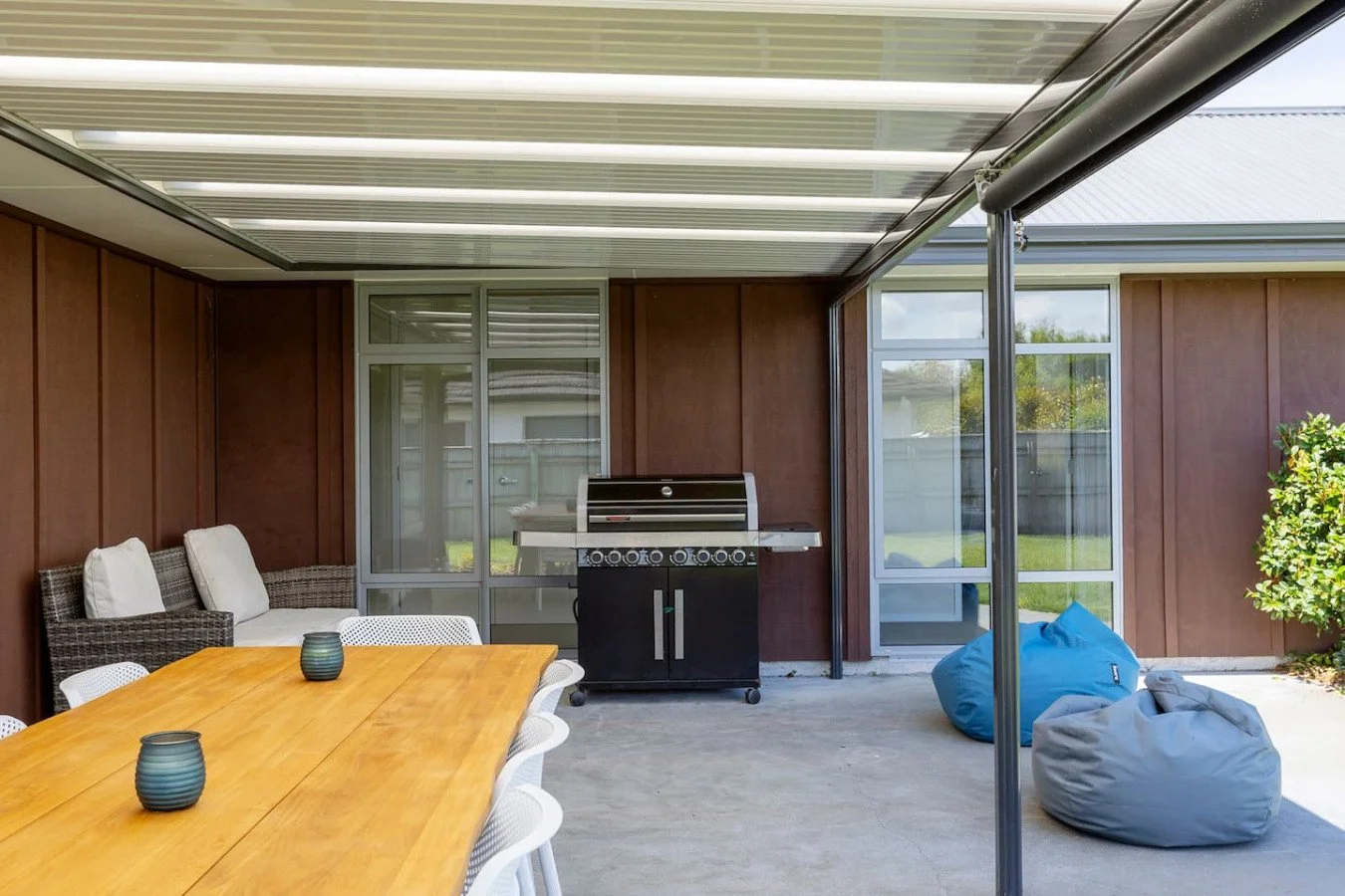 An outdoor patio with a wooden table, wicker and white chairs, a black barbecue grill, two bean bags, and a screened enclosure with sliding glass doors and windows, surrounded by a fence and greenery.