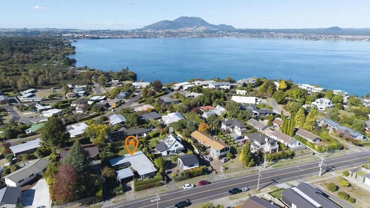 Aerial view of a residential neighborhood near a body of water with a mountain in the background. Several houses with trees and greenery surround the area, and a main road runs across the bottom of the image.