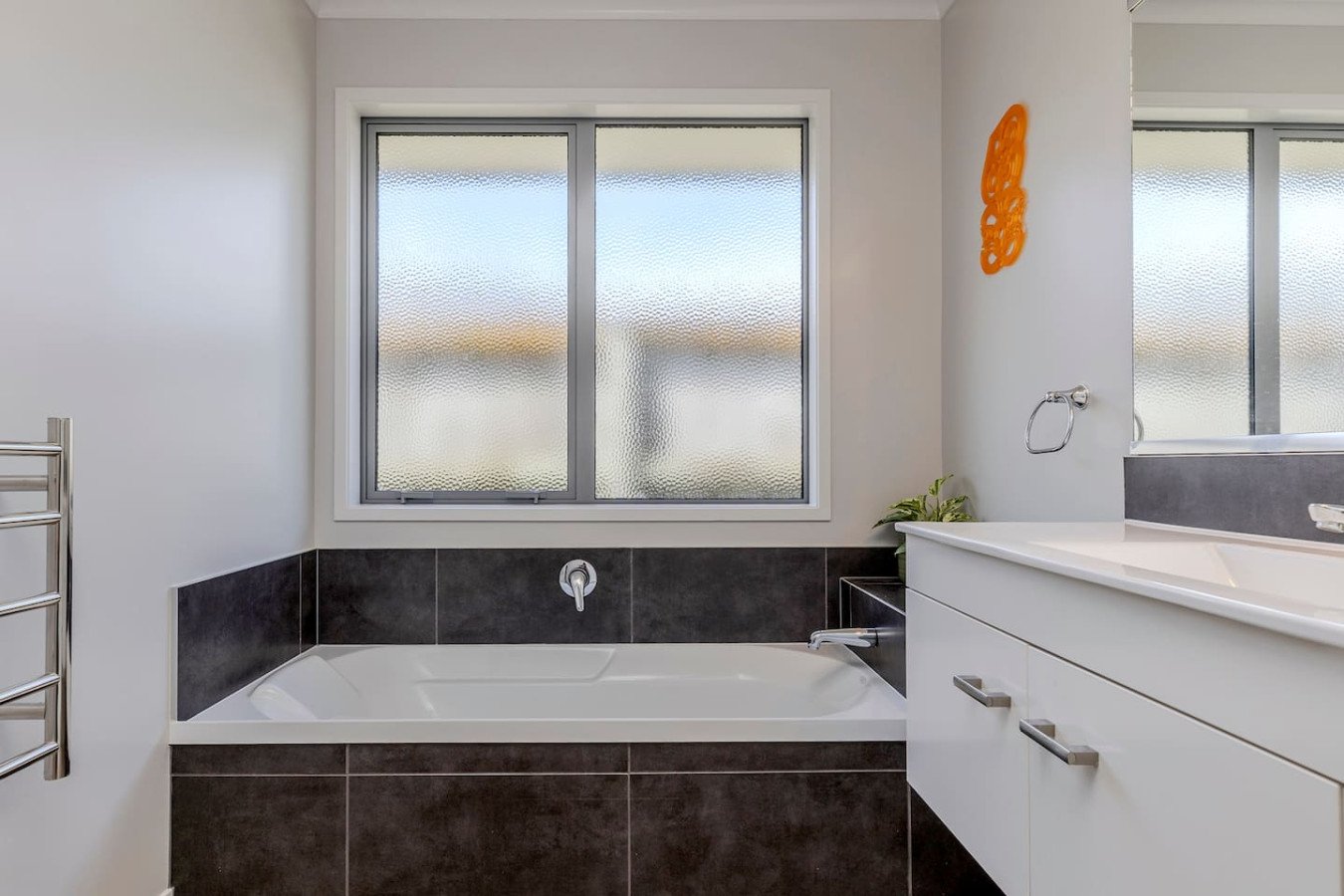Modern bathroom with a bathtub under a frosted window, dark tile around the tub, white vanity with drawers, and a wall-mounted towel ring.