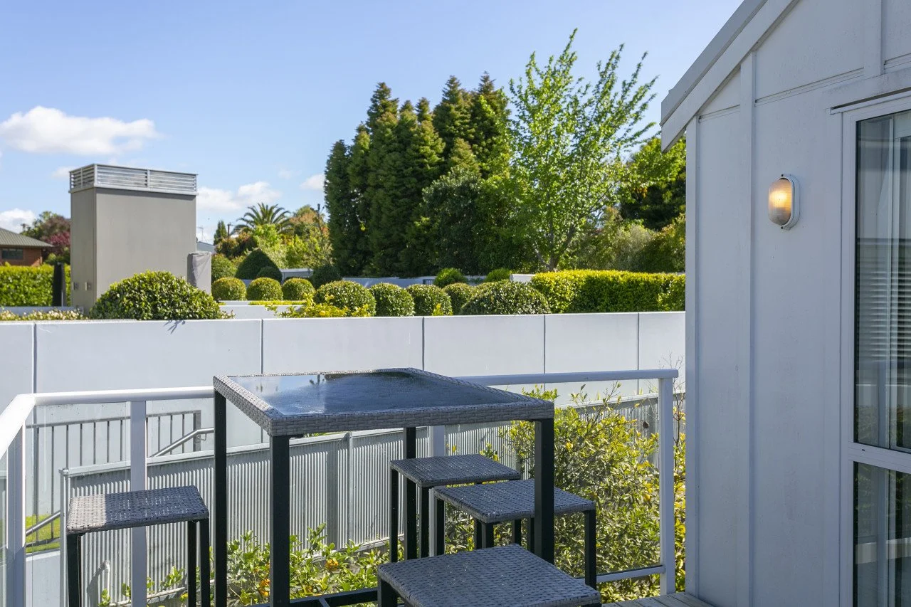 Small balcony with black wicker table and chairs, white railing, white building wall with window and outdoor light fixture, lush green trees and bushes in the background, clear blue sky.