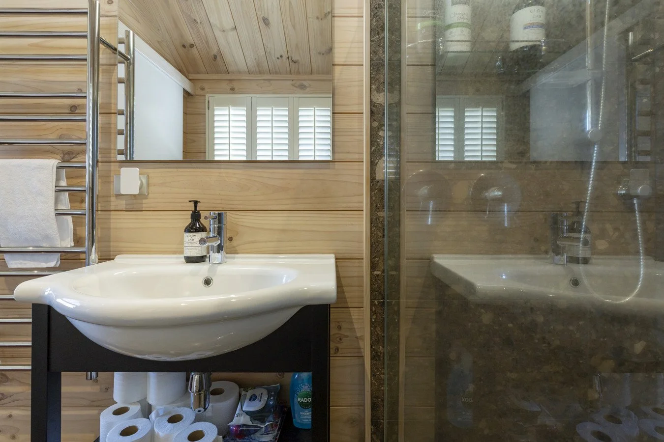 Bathroom with a wooden wall, a white sink with a black base and toiletries on it, a mirror, a towel on a metallic rack, and a shower with a glass door. Reflection in the mirror shows a window with shutters.