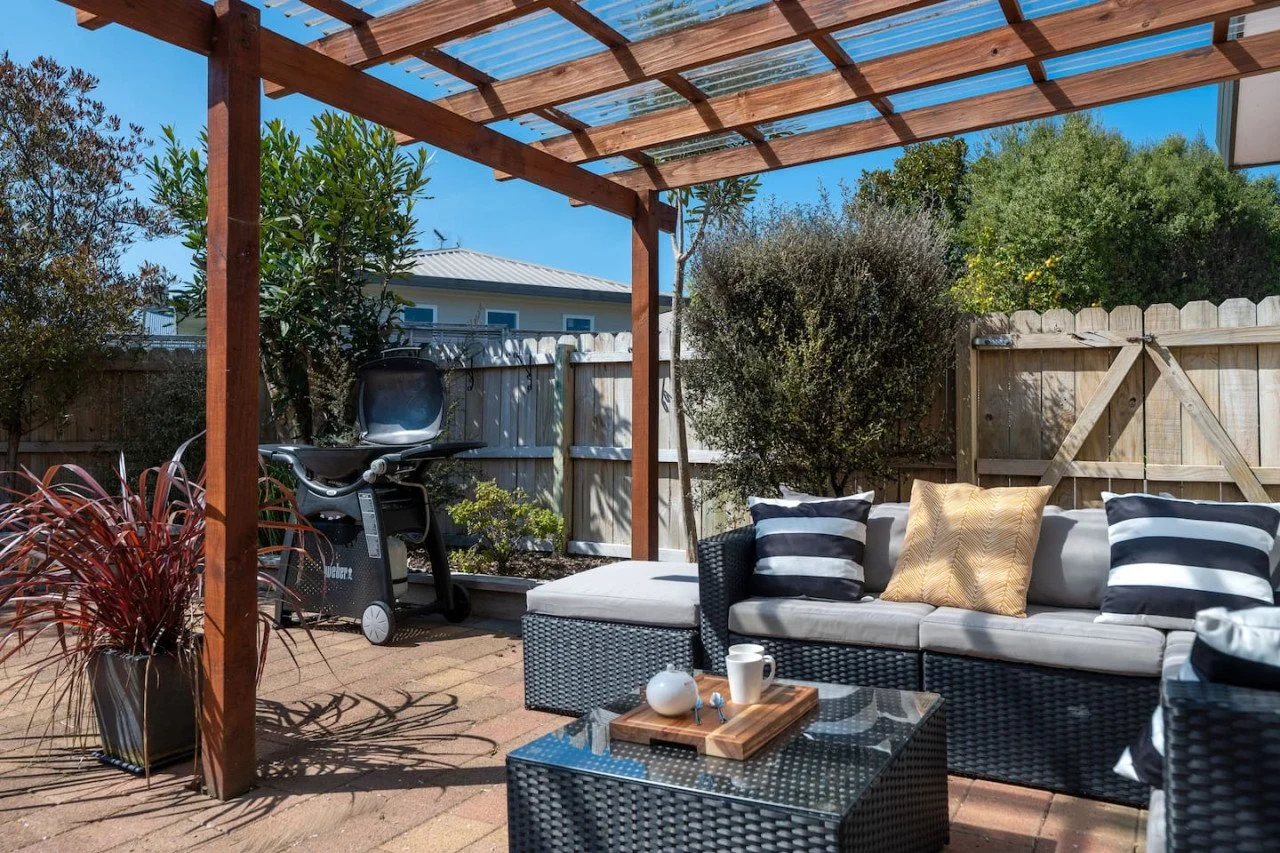 Outdoor patio with a woven sofa with striped and solid-colored throw pillows, a glass-topped coffee table with a wooden tray and cups, a barbecue grill, potted plants, and a wooden pergola under a blue sky.