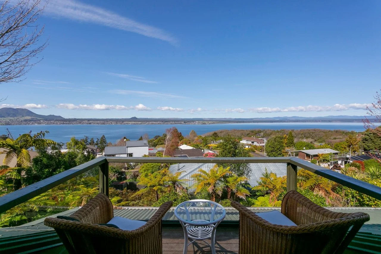 View from a balcony overlooking a lake surrounded by trees and houses, with mountains in the distance and a clear blue sky.