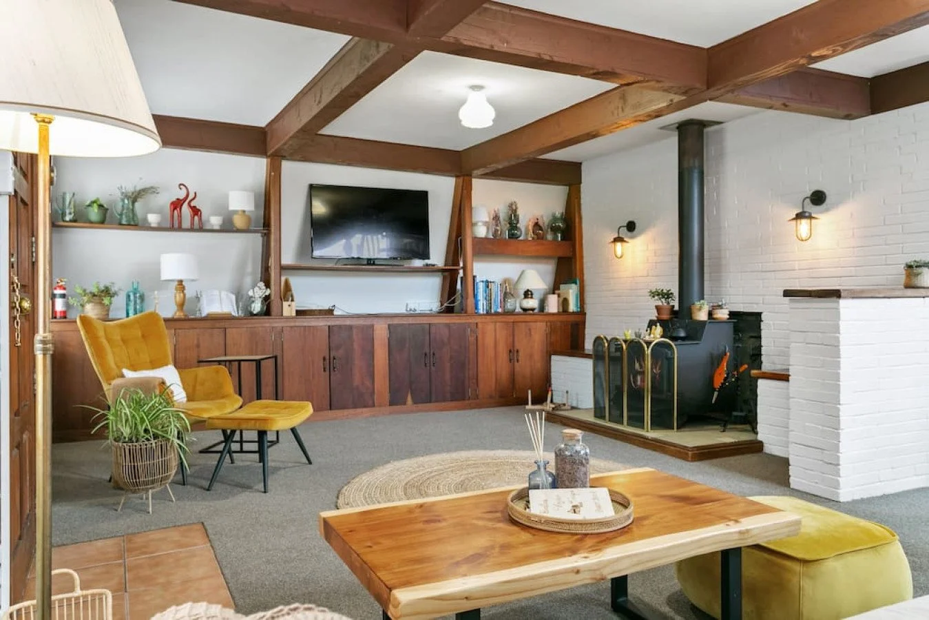 Living room with wooden beams, a fireplace, TV, yellow chair, houseplants, decorative shelf, wooden coffee table, and beige carpet.