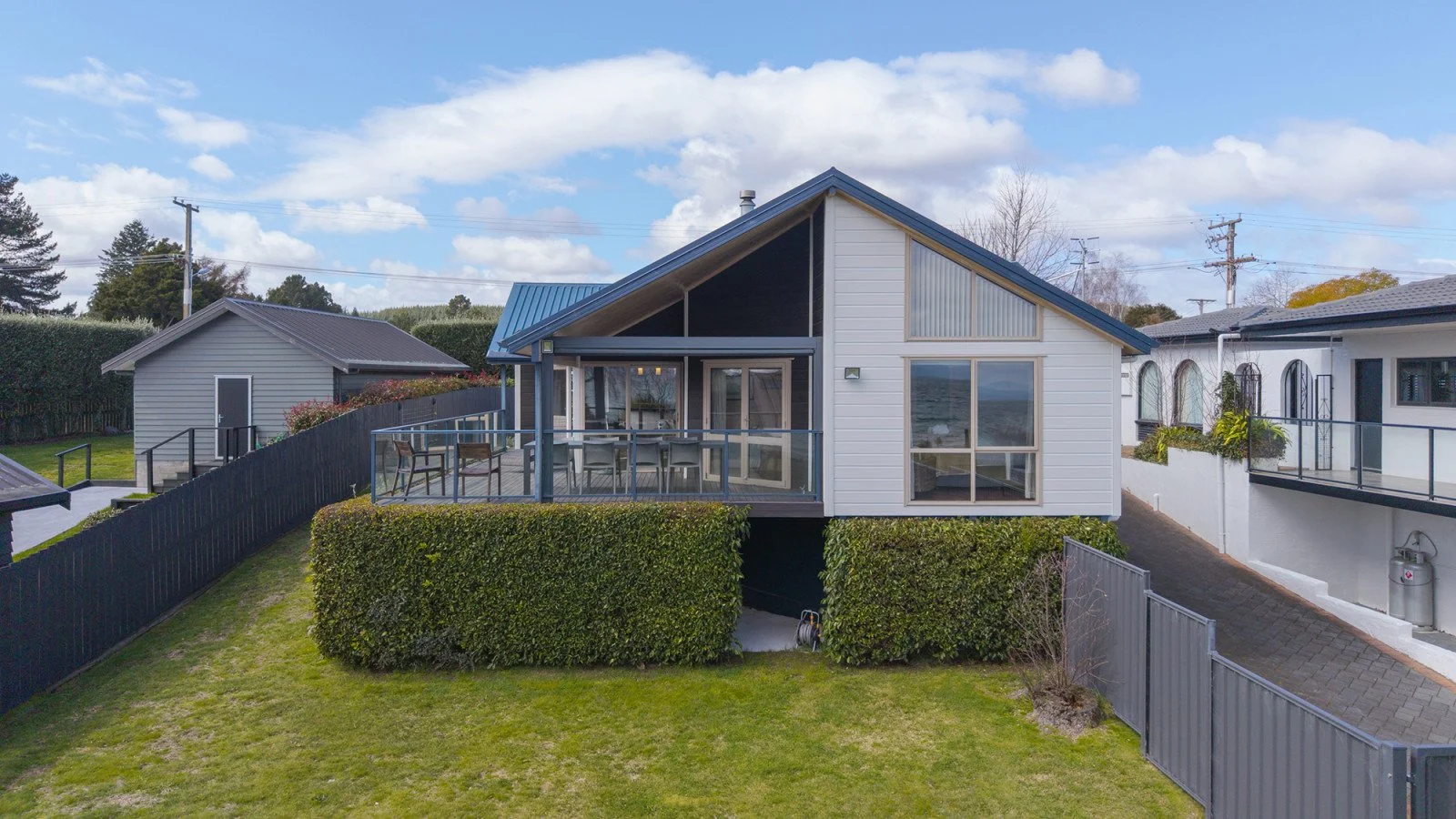A modern house with a large balcony, surrounded by a well-maintained lawn and neighboring houses, under a partly cloudy sky.