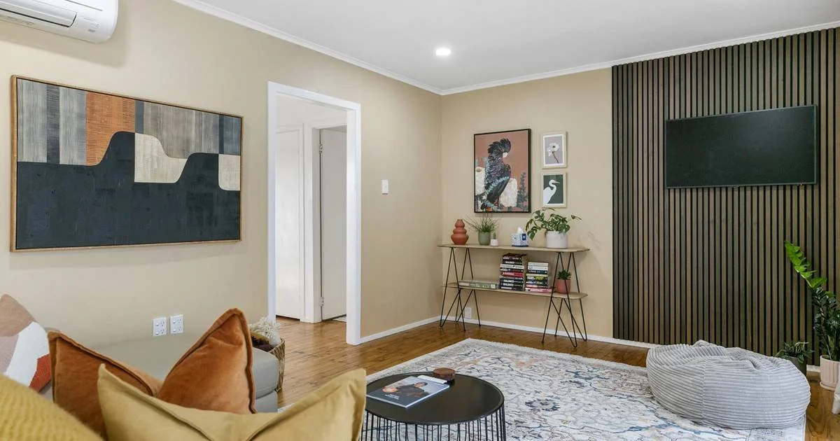 Living room with beige walls, wooden flooring, a black accent wall with wood slats, a wall-mounted flat-screen TV, framed artwork, a bookshelf, a beige sofa with colorful pillows, a rug, a round coffee table, a bean bag, and potted plants.