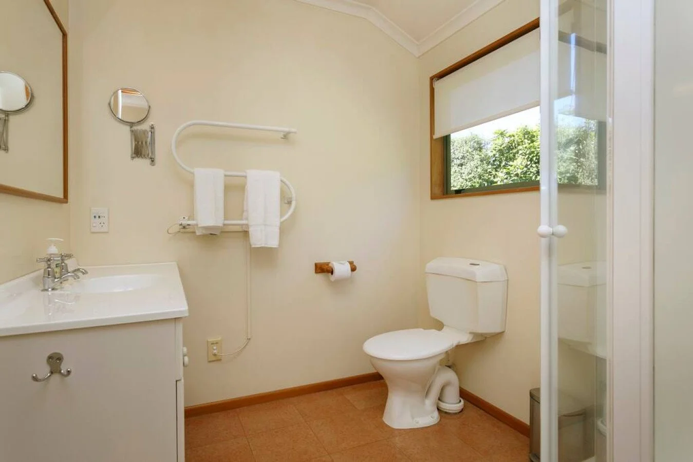 A small bathroom with a white toilet, a small white sink with a silver faucet, a mirror, a window with a view of green trees, and a grab bar with towels. There is a shower enclosure in the right corner.