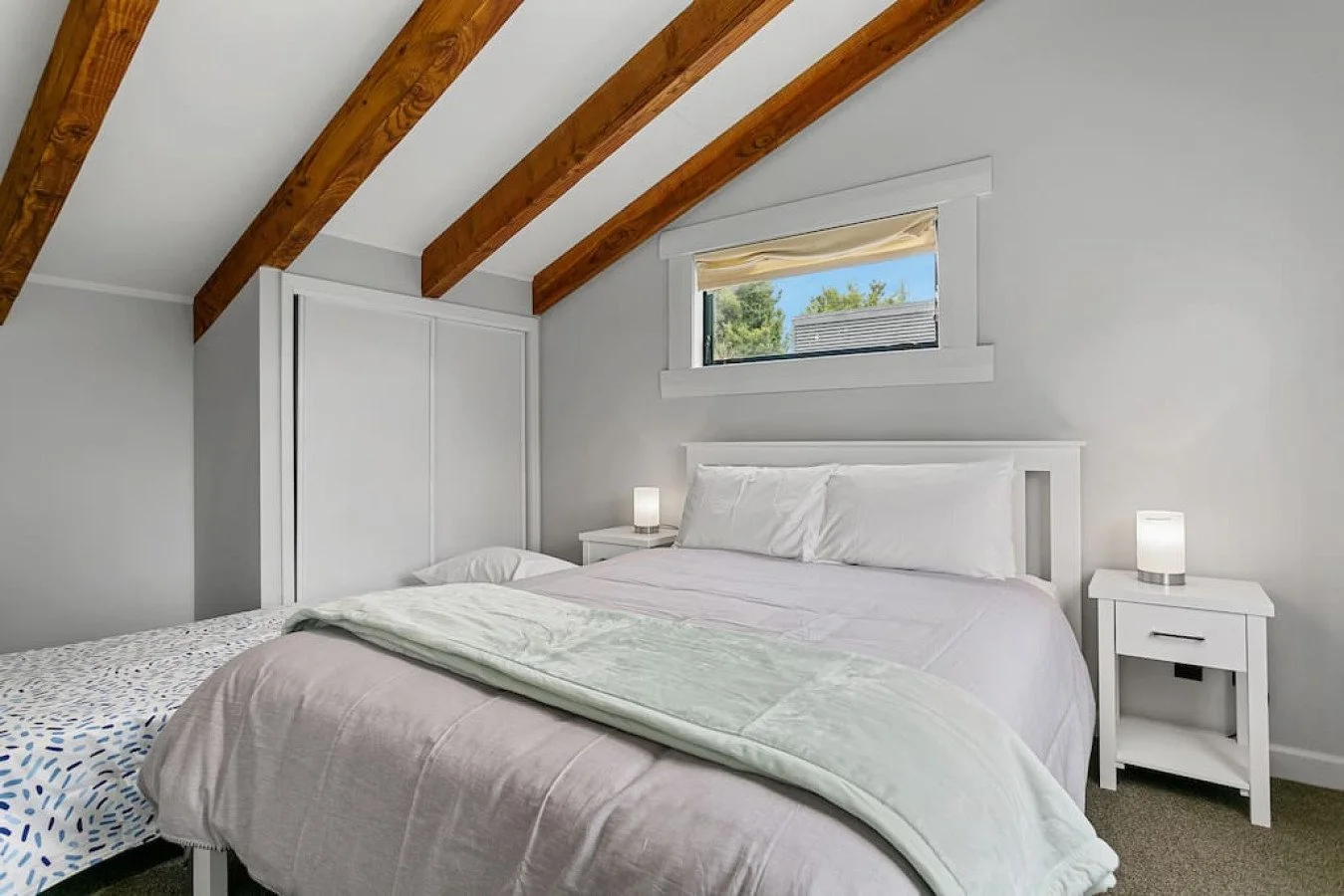 A bedroom with a vaulted ceiling and exposed wooden beams, featuring a white bed with white pillows and comforter, two white nightstands with lamps, a window with a beige shade, and a view of trees outside