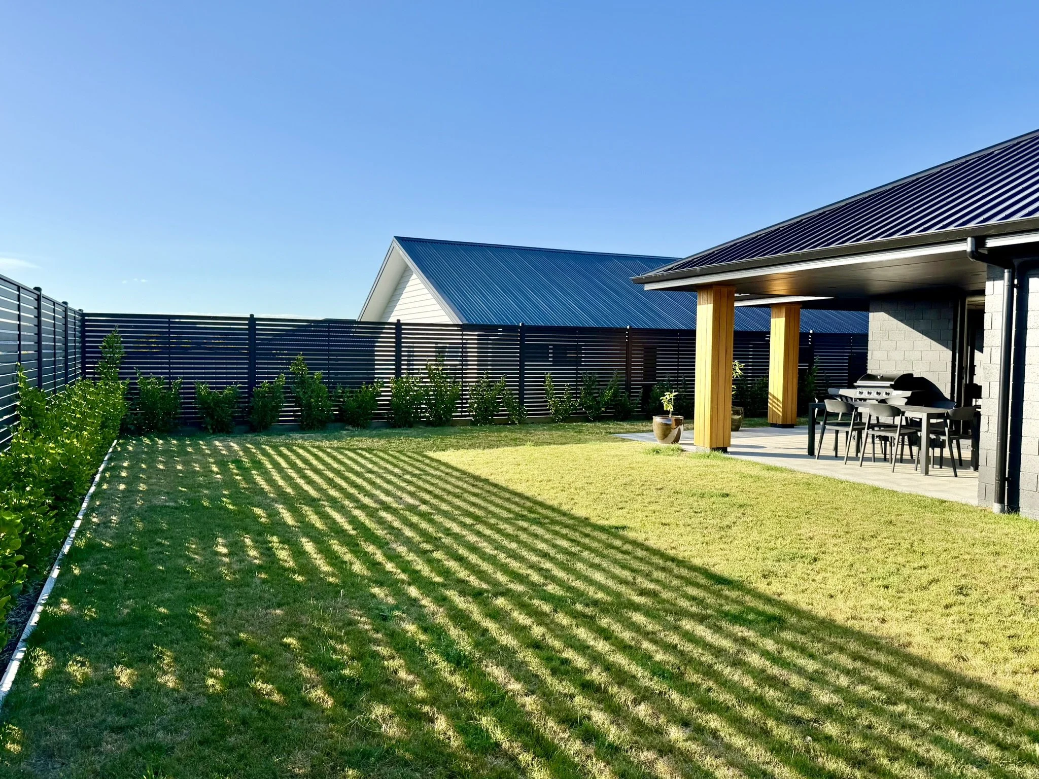View of a backyard with a grassy lawn, a black metal fence, some green shrubs, and a covered patio with a patio table, chairs, a grill, and potted plants. There are houses with blue and dark gray metal roofs in the background under a clear blue sky.