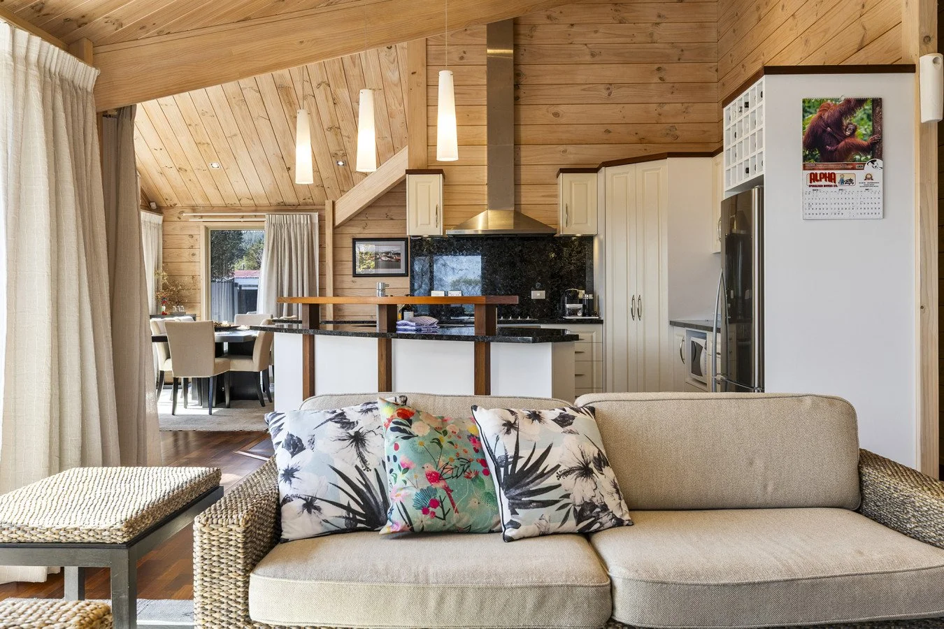 Open concept living room and kitchen with wood-paneled walls and ceiling, beige sofa with floral pillows, kitchen with white cabinets, black granite countertops, stainless steel appliances, and a dining area with a round table and beige chairs.