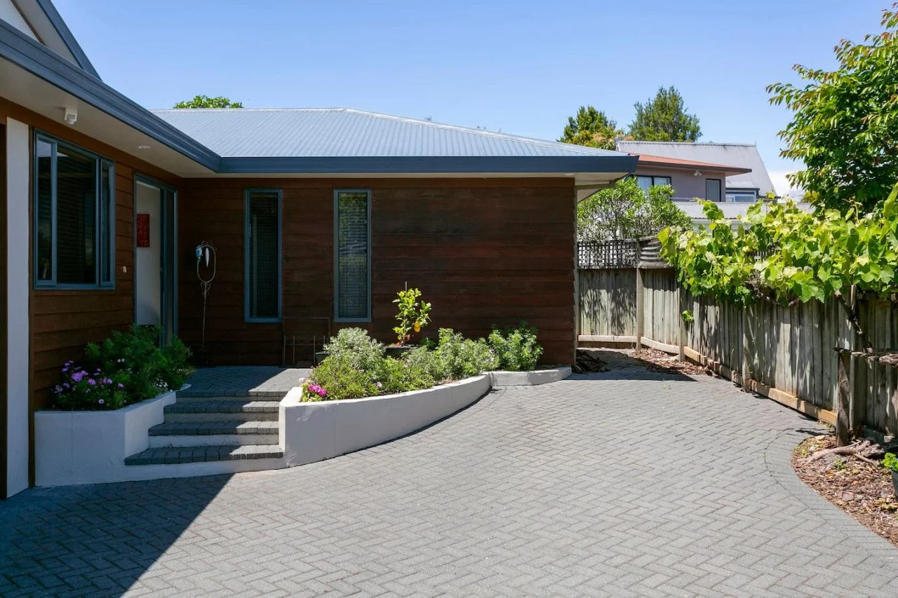 A modern house with wooden exterior walls, a metal roof, and a paved driveway. There are small garden beds with plants and flowers, a white curved retaining wall, and a wooden fence with greenery along the edge. The sky is clear and blue.