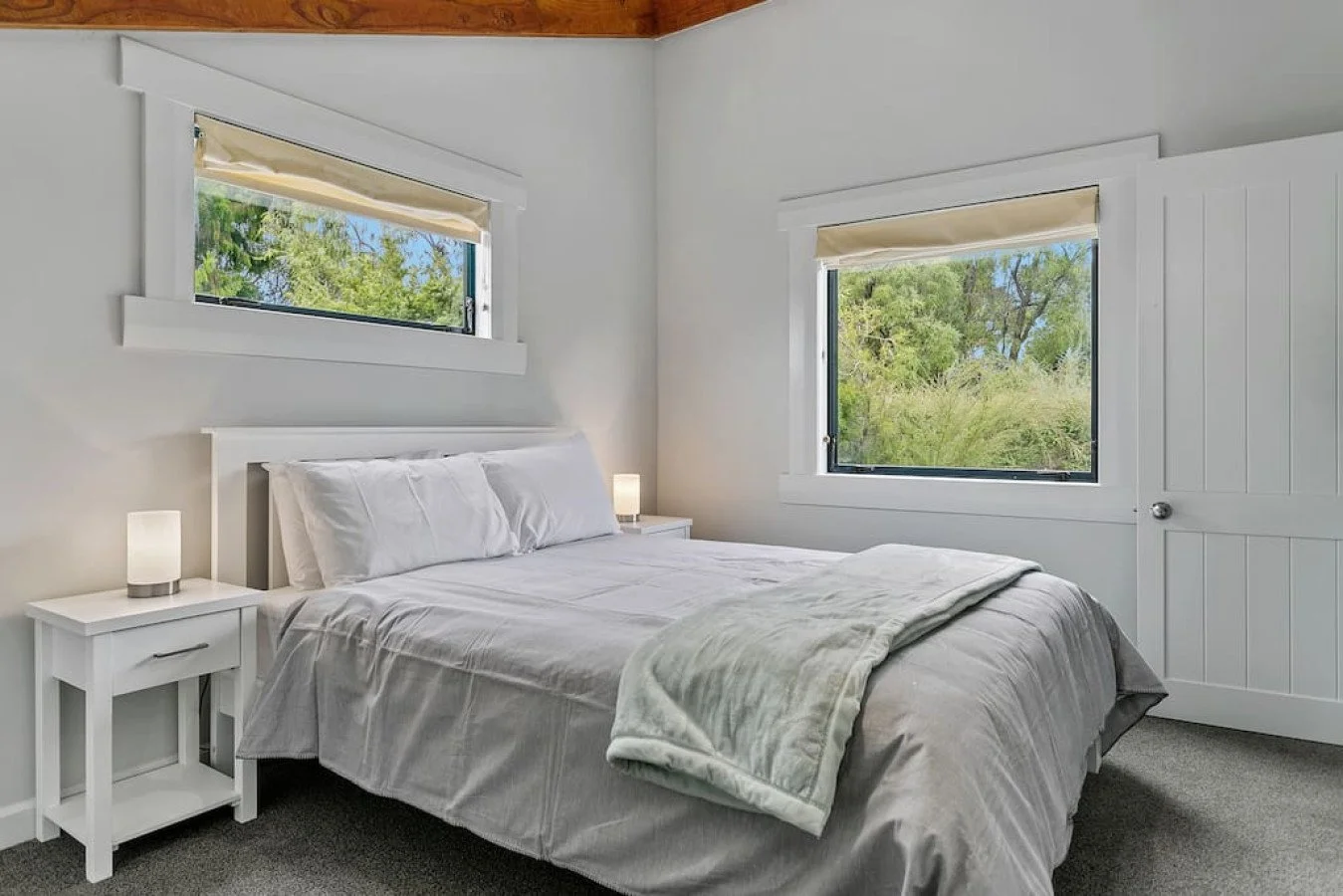 A cozy bedroom with a bed featuring white bedding and pillows, two white nightstands with lamps, and two large windows showing green trees outside.
