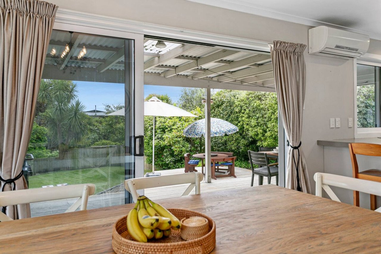View from kitchen into a backyard with a wooden table, bananas in a basket, and an outdoor patio with umbrellas and garden.