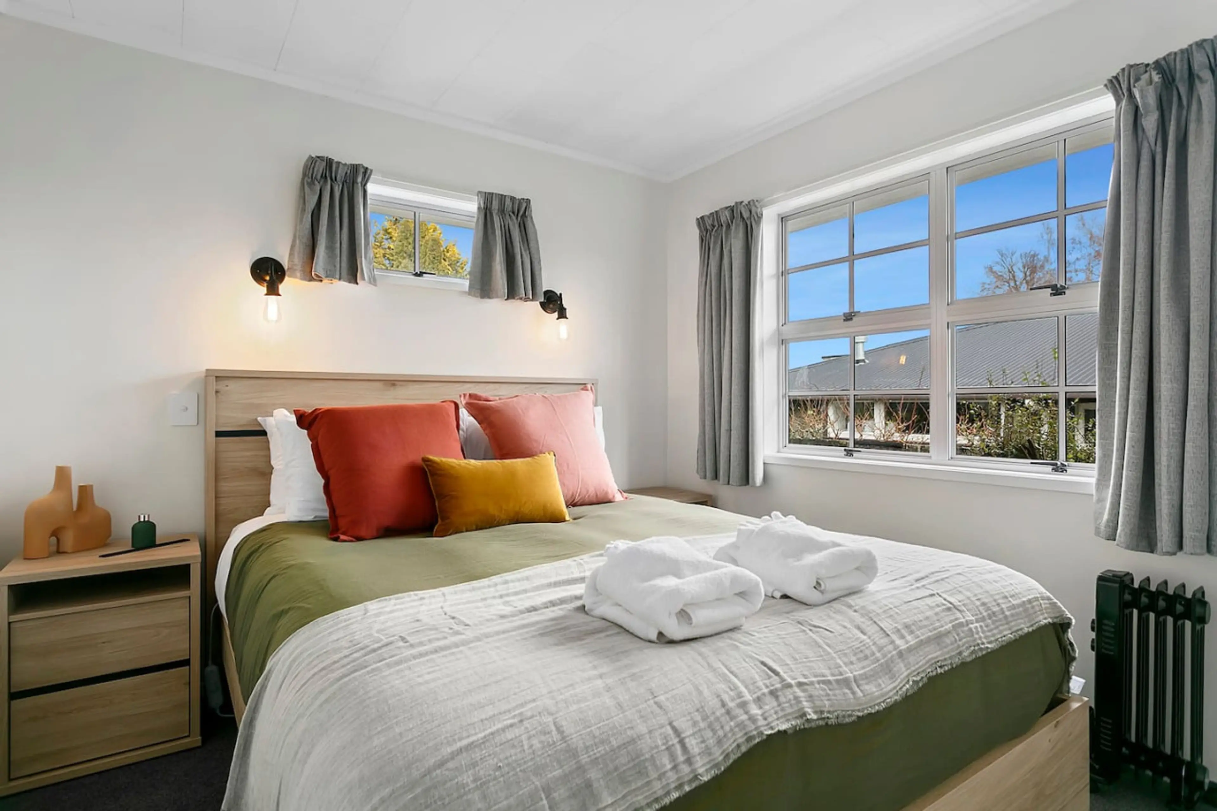 A neatly made bed with a green bedspread, colorful pillows, and towels on top in a bright bedroom. There are two windows with gray curtains, and wall-mounted lights above the headboard. A small wooden nightstand with decorative items and a black radi
