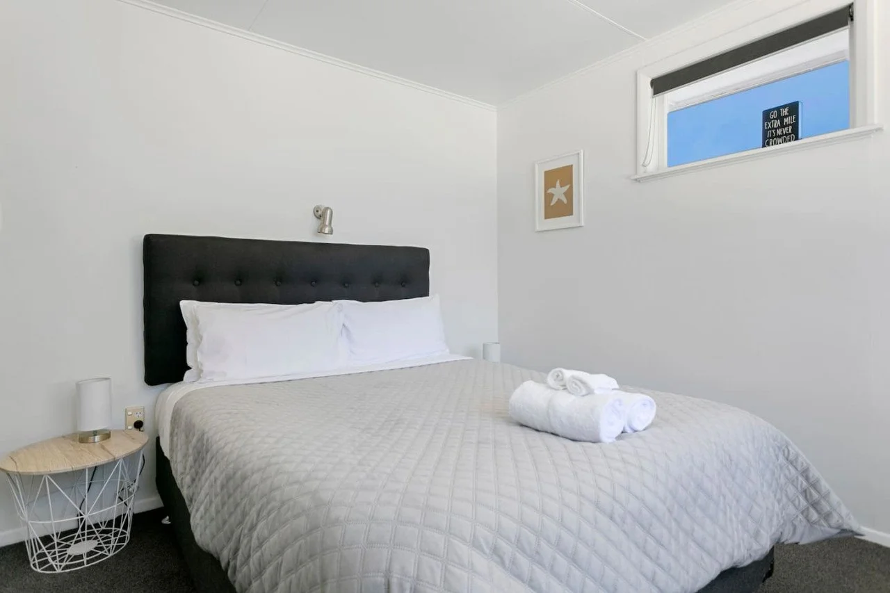 Clean bedroom with a double bed, white bedding, folded towels on the bed, dark upholstered headboard, small round bedside table with lamp, framed starfish artwork on the wall, and a window with a sign outside that says "Go the extra mile. It’s never 