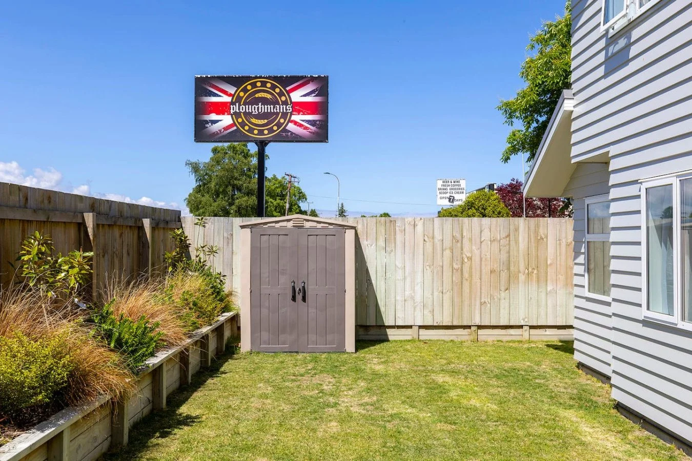 A backyard with a wooden fence, a gray garden shed, a small garden bed with plants, a house with white siding, and a billboard with a British flag and the text 'ploughmans' in the background.