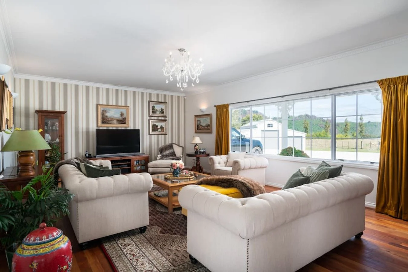 Bright living room with white sofas, wooden coffee table, large window with yellow curtains, and a chandelier