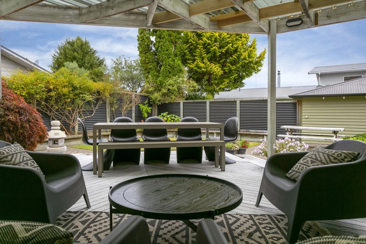 Outdoor patio with seating, including armchairs, a coffee table, and a dining table with chairs, covered by a wooden pergola. The patio is enclosed by a fence with trees and plants in the background.