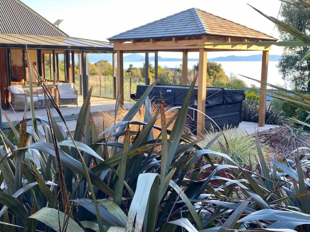 Outdoor deck overlooking water with a hot tub under a wooden gazebo and seating area with chairs and a table, surrounded by garden plants.