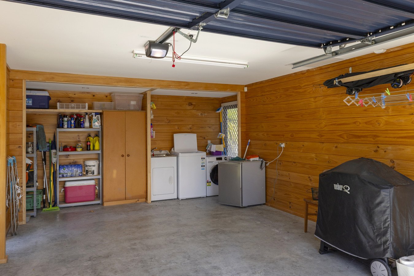 Empty garage with wooden walls, concrete floor, washing machine, dryer, shelves with cleaning supplies, a covered barbeque grill, and hanging laundry clips.