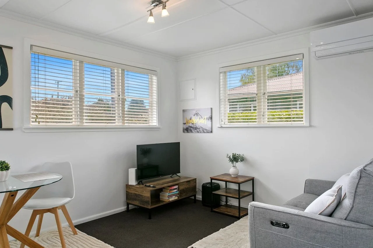 Living room with white walls, large windows with blinds, a gray sofa, a small wooden TV stand with a television, a small table with a potted plant, and a minimalist design.