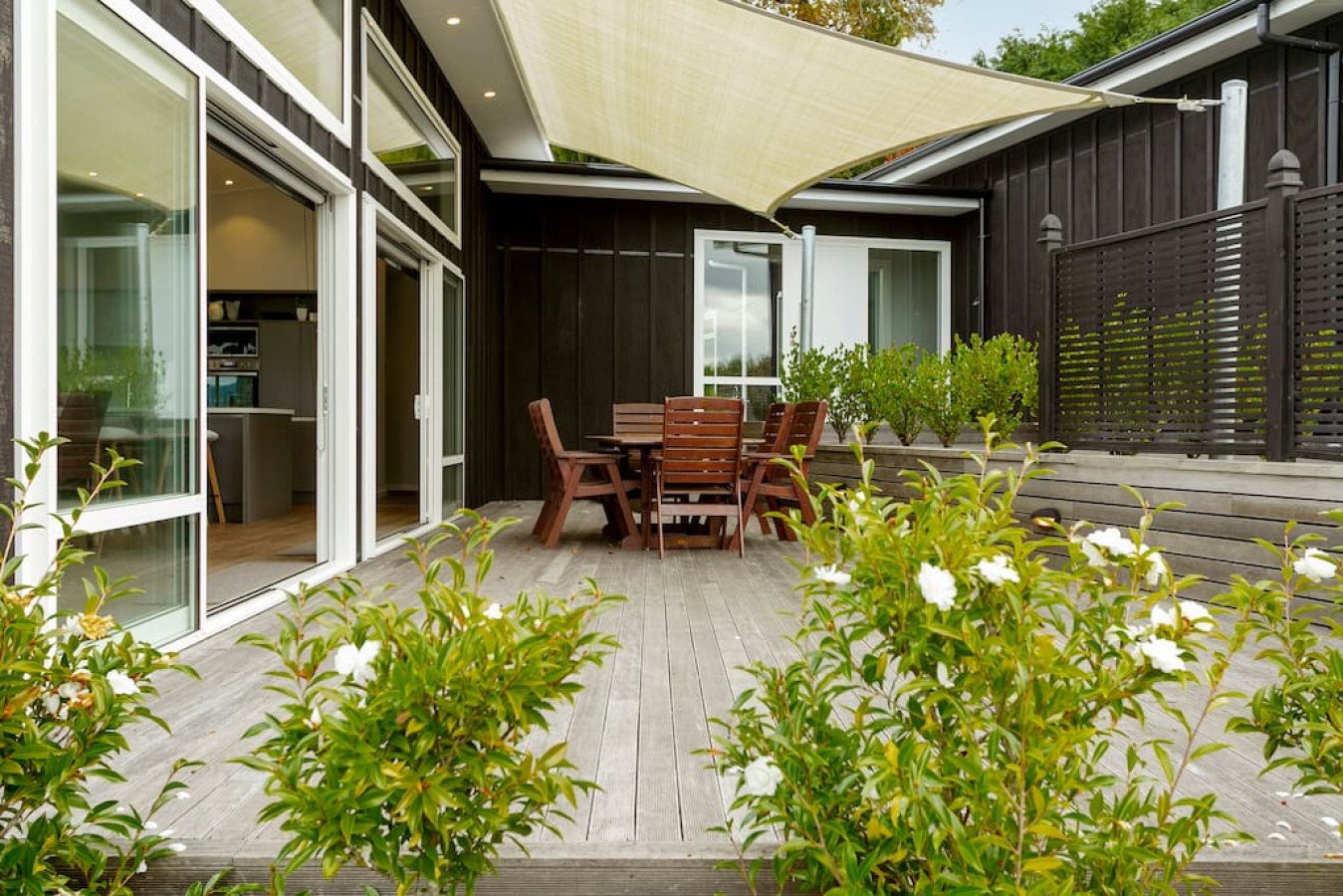 Outdoor patio with wooden deck, brown dining table with six chairs, white flowering bushes, black wooden house wall, sliding glass doors, and beige fabric shade sail