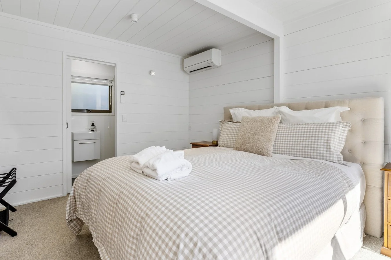 A neatly made bed with beige and white checkered bedding, several pillows, and a folded towel on top, in a bright, minimalistic white bedroom with an air conditioning unit.