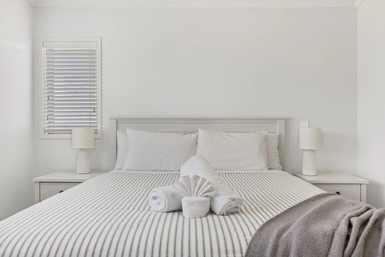 A neatly made bed with white pillows and striped bedding, flanked by two white bedside tables with lamps, in a bright and minimalist bedroom.