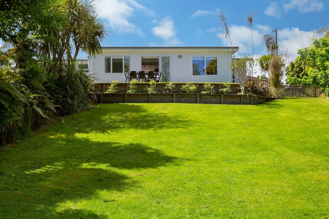 A backyard with a grassy lawn, trees, and plants, leading up to a raised wooden deck attached to a white house with sliding glass doors and windows.