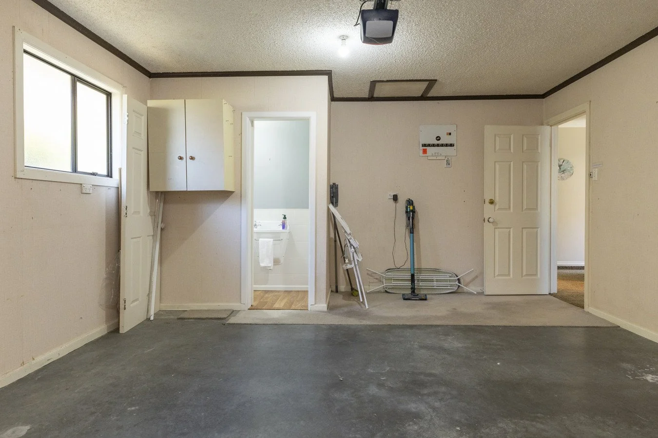 Empty room with a concrete floor, white walls, a window on the left, and a ceiling with a hatch. There are objects leaning against the walls, including a folded drying rack and a cordless drill. An open door reveals a small bathroom with a sink and t