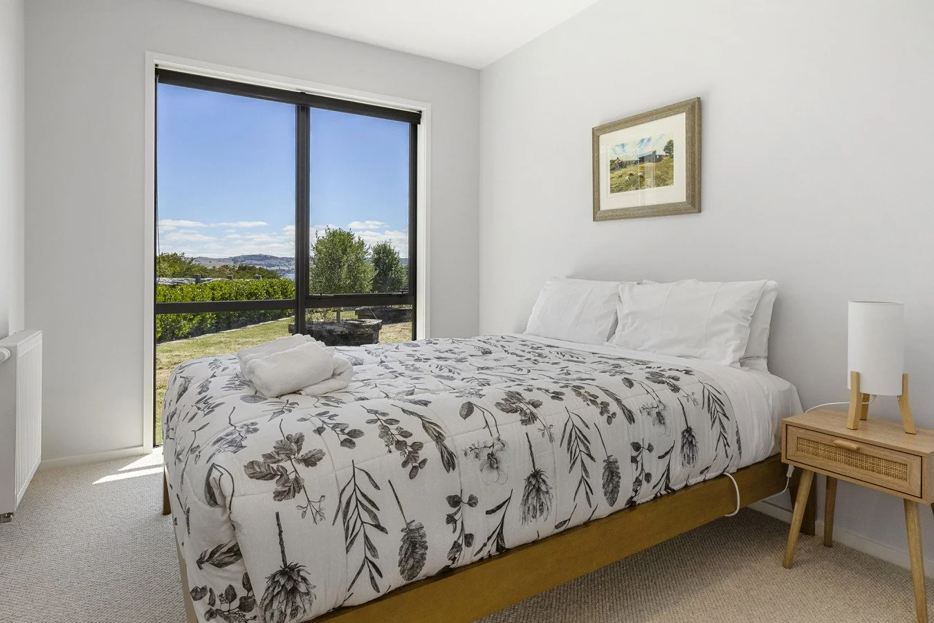 Bedroom with a bed covered in white and gray floral duvet, pillow, and a folded towel, next to a wooden nightstand with a lamp, and a large window showing an outdoor view of trees, grass, and hills.