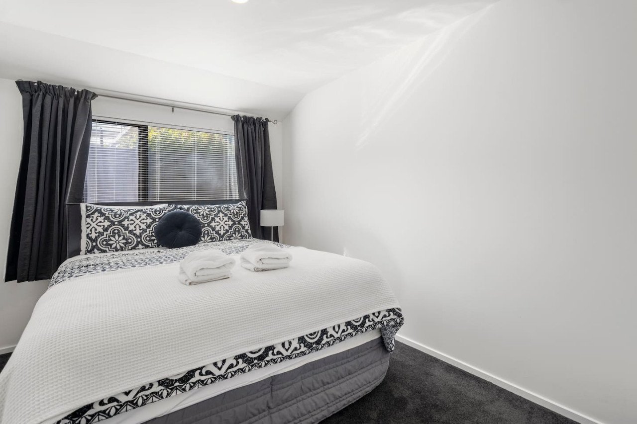 A neatly made bed with black and white patterned pillows and a white textured blanket, two folded towels placed on top, in a minimalist bedroom with dark curtains and a small white nightstand next to a window.