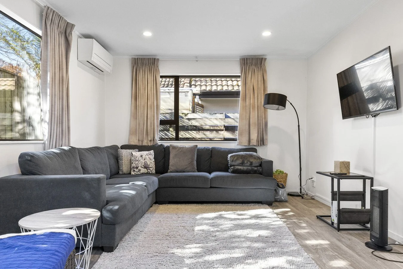 Living room with a large gray sectional sofa, a small white round side table, beige curtains, a window showing an outside fence and roof, a black floor lamp, a wall-mounted TV, a small table with a beige speaker, and a black space heater.