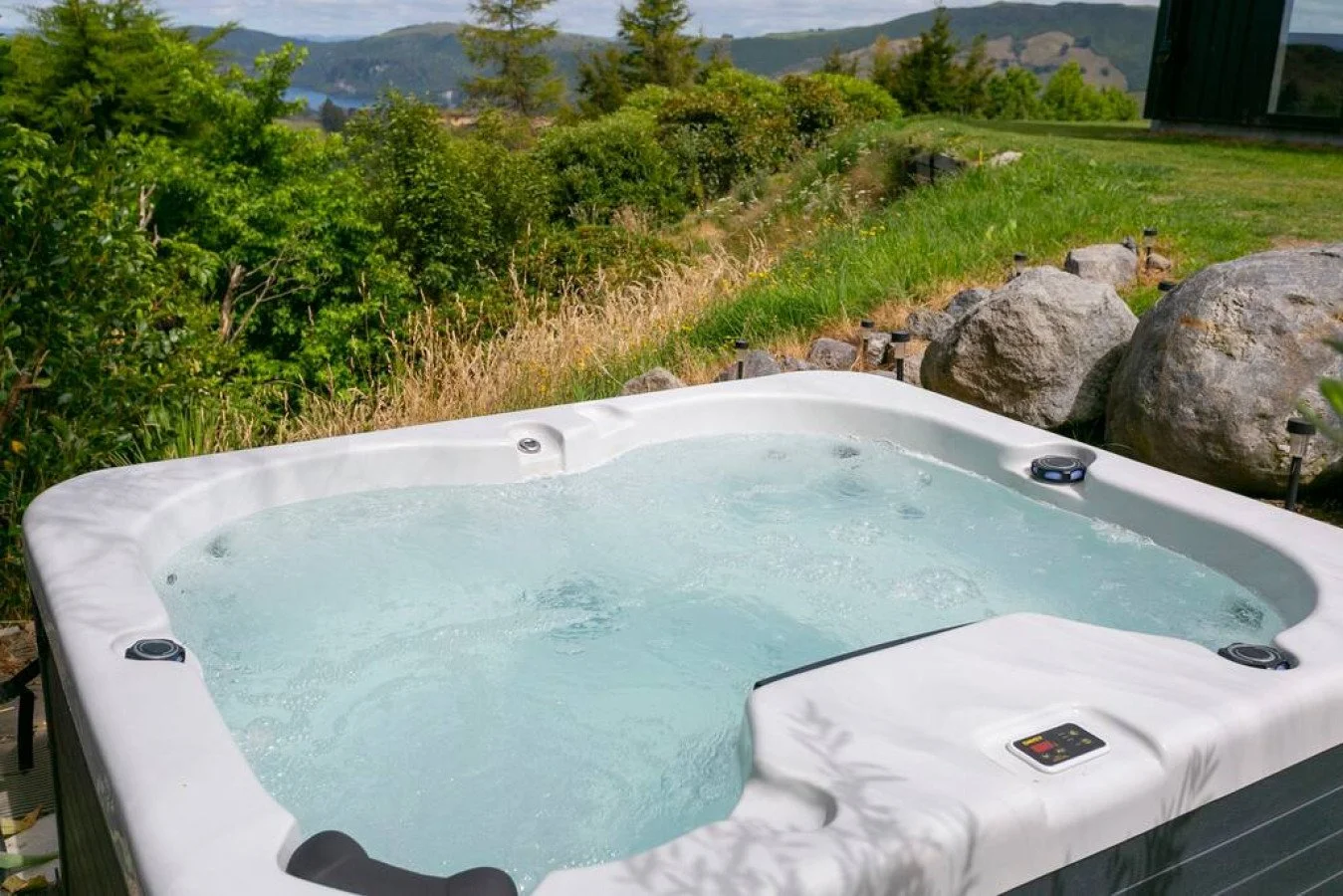An outdoor hot tub filled with bubbling water, set in a grassy area with rocks and lush green trees, overlooking a scenic landscape of hills and mountains.
