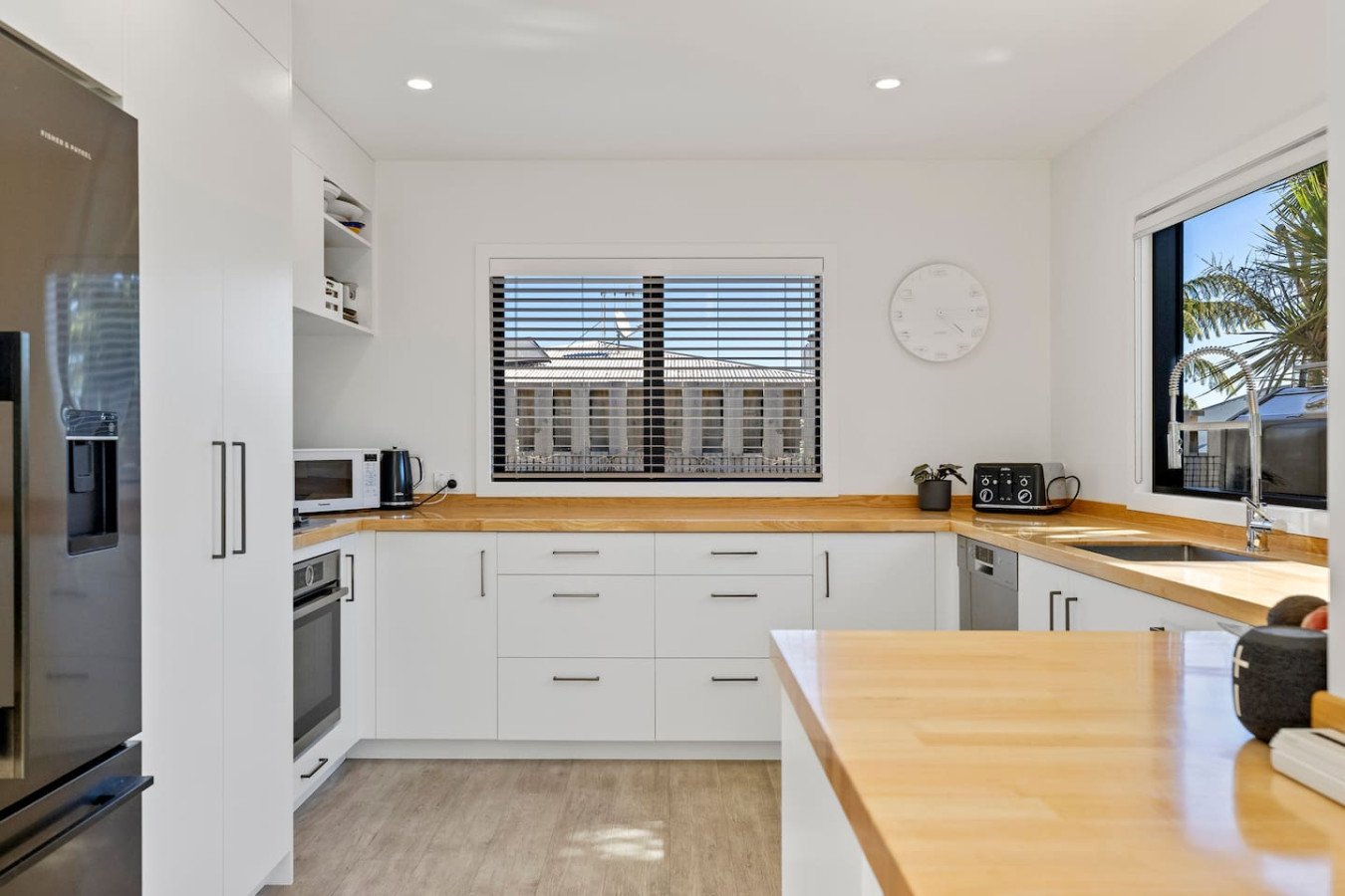 Bright, modern kitchen with white cabinets, wooden countertops, stainless steel appliances, and large windows showing a building and palm tree outside.