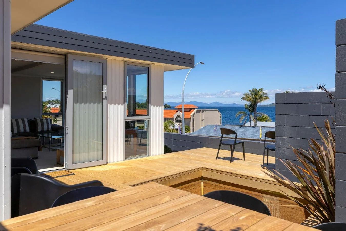 Outdoor patio with wooden flooring, black chairs, a view of the ocean, palm trees, and neighboring buildings with red and gray roofs under a clear blue sky.