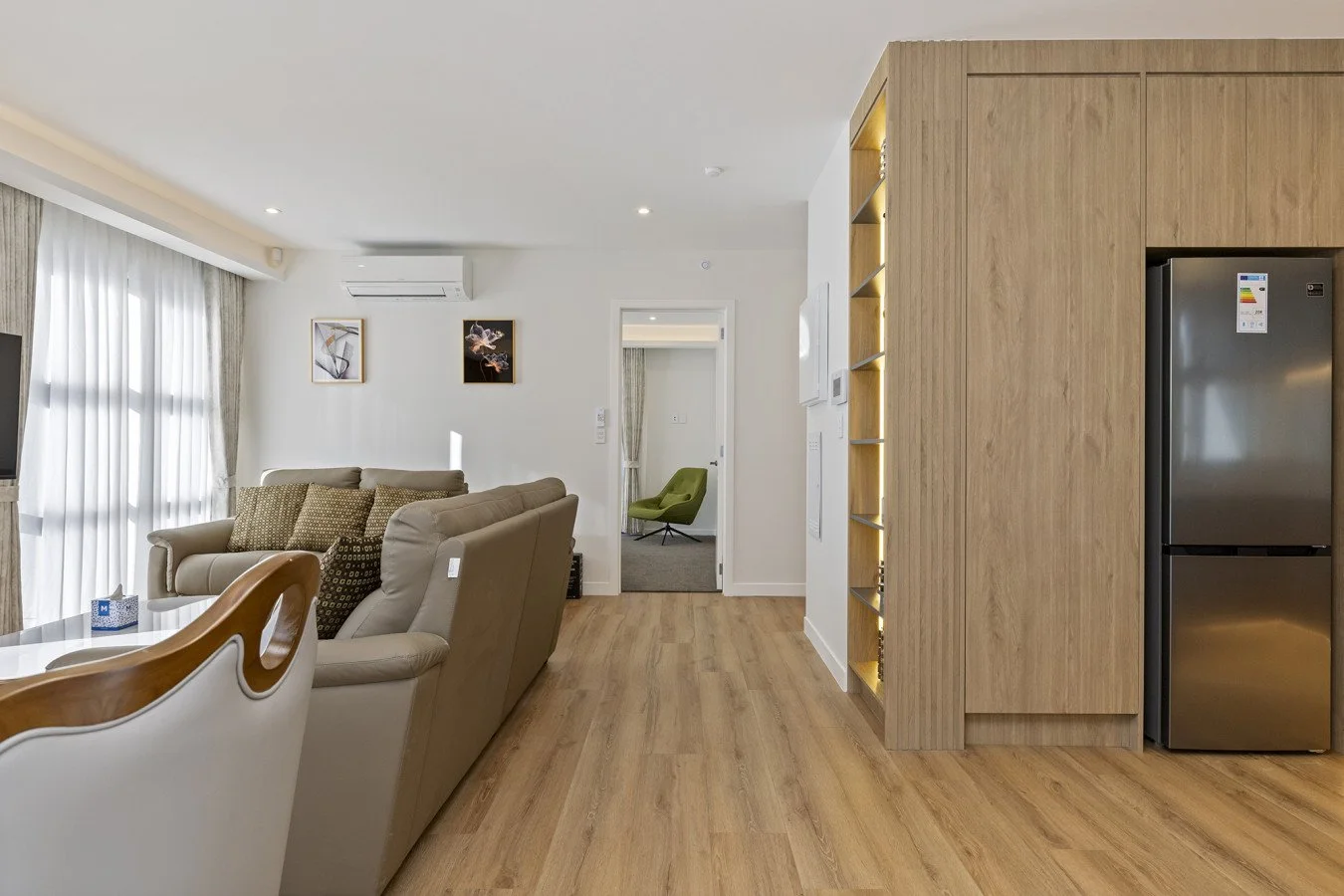 Modern living room with beige sofas, a dining table, a large window with curtains, wall art, an air conditioner, and hardwood floors, with a doorway leading to a room with a green chair and gray carpet.