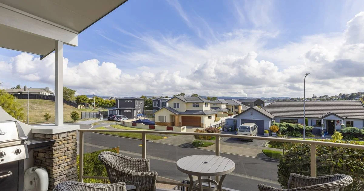 View from a balcony overlooking a suburban neighborhood with houses, parked cars, and a street under a partly cloudy sky.