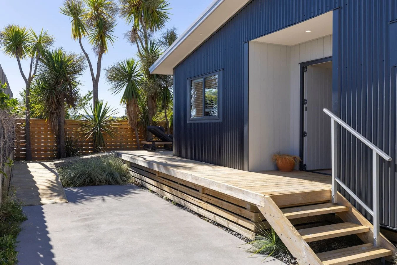 Front porch of a modern house with blue metal siding, wooden steps, and a white railing. There is a small potted plant near the door, a window, and desert plants with tall trees in the background.