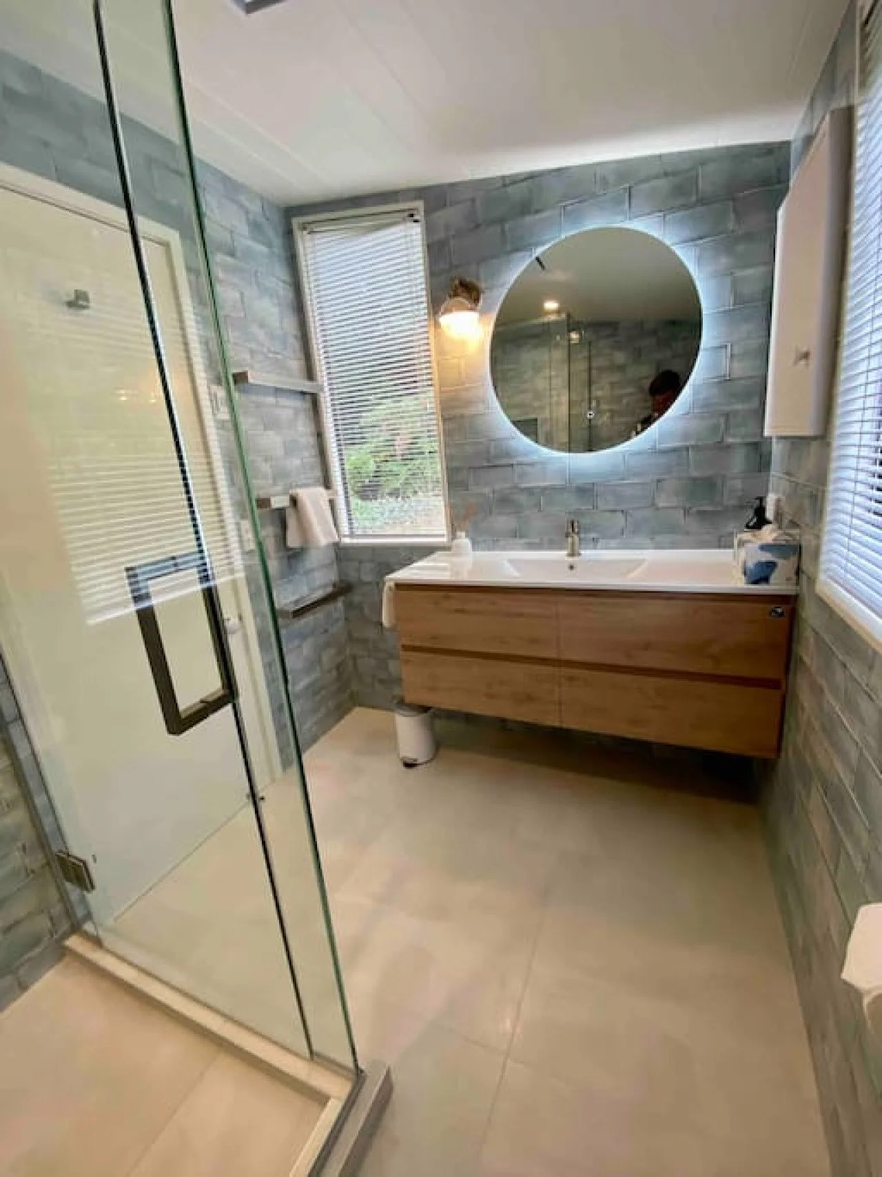 Modern bathroom with brick-patterned gray tiles, a glass shower enclosure, a wooden vanity with a white sink, a large round mirror, and windows with blinds.