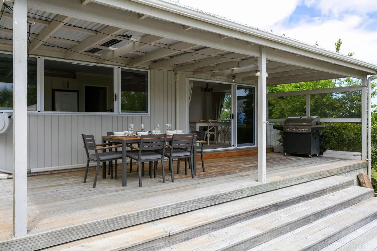 Outdoor patio with a wooden dining table, six chairs, a barbecue grill, and a sliding glass door leading to the interior of a house, surrounded by trees.