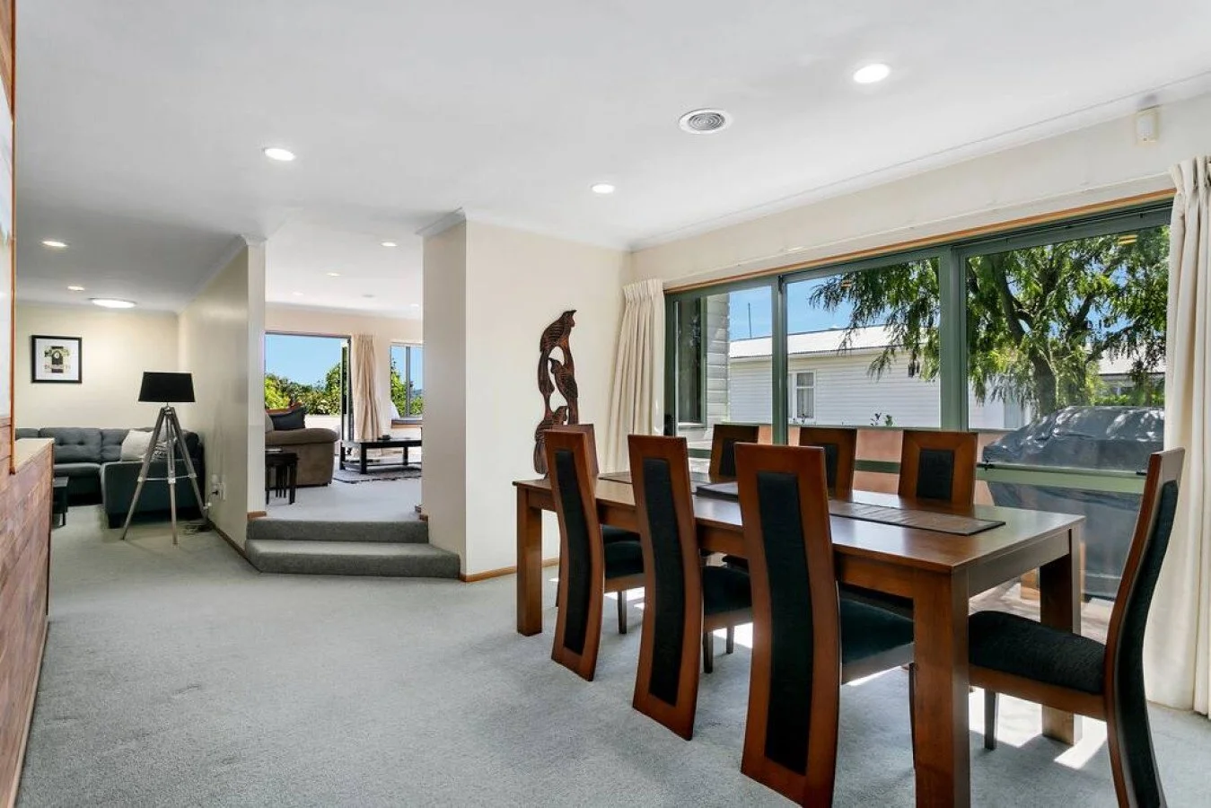 Dining room with large windows, six wooden chairs with black cushions around a wooden table, and a carved wooden statue on a side table. Adjacent living room with gray couch, black floor lamp, and a doorway leading outside.