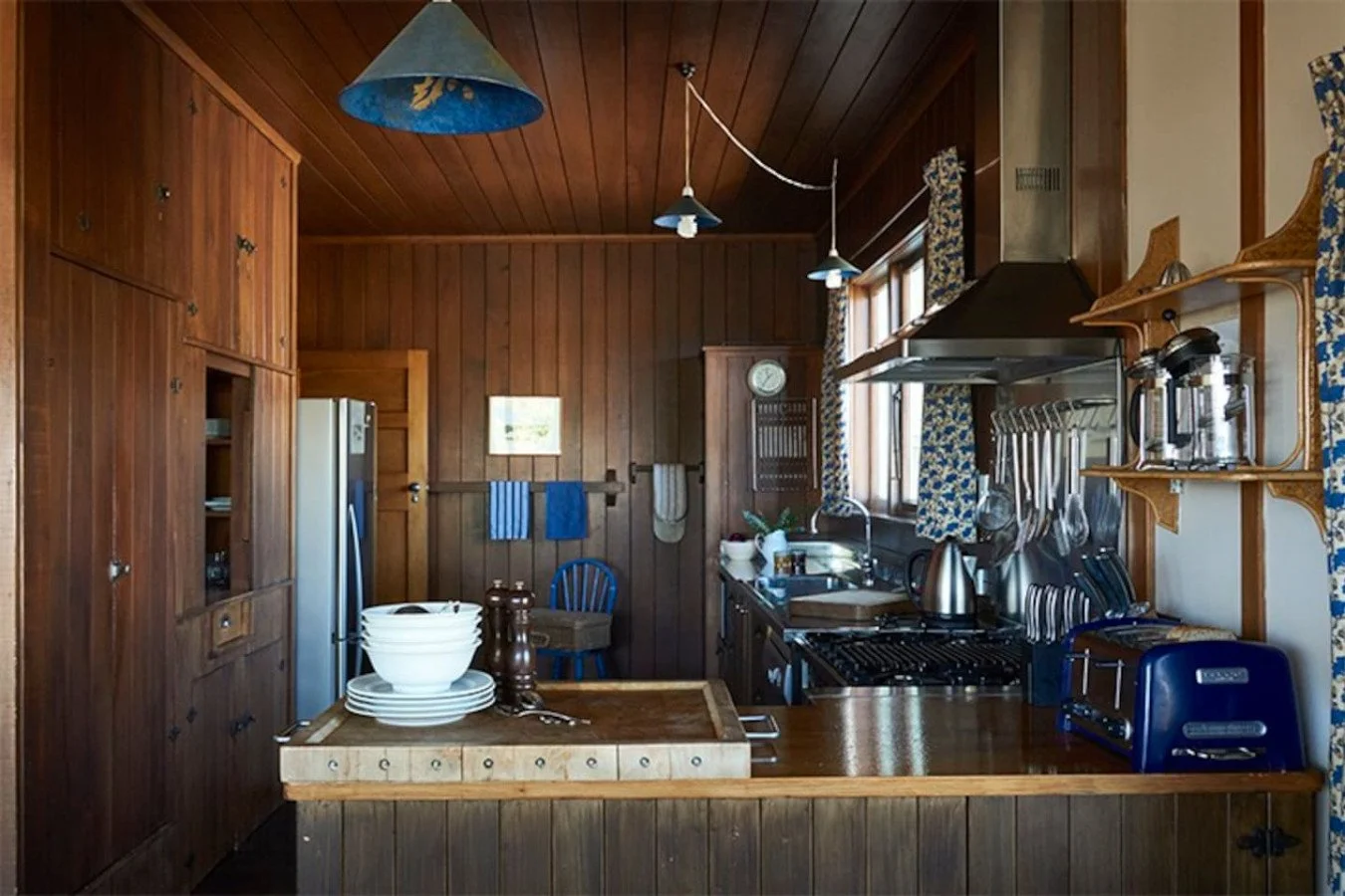 Rustic kitchen with wooden cabinets, a stainless steel refrigerator, a stove, and a wooden island with bowls and salt and pepper shakers. There are hanging blue lamps, patterned curtains, and kitchen utensils on a rack.