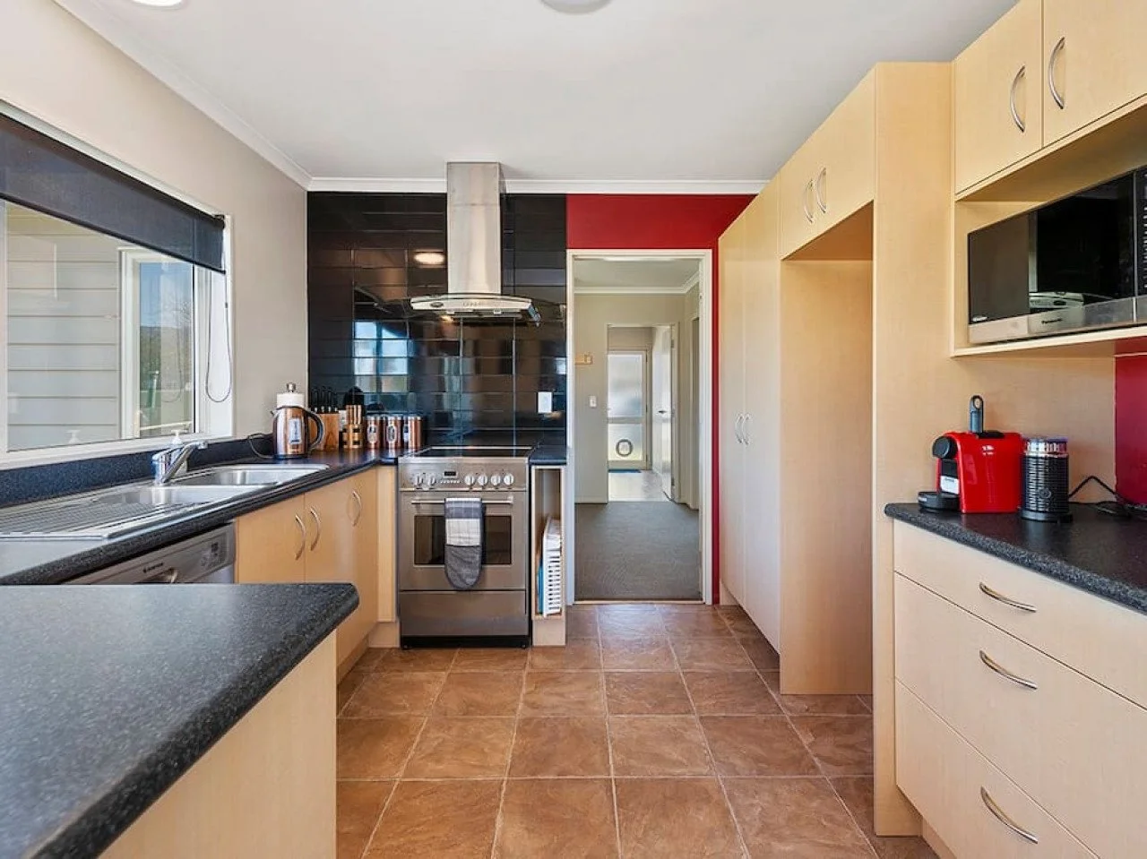 A modern kitchen with beige cabinets, black countertops, and a tiled floor. There is a window with a black blind, a stainless steel stove, and various kitchen appliances, including a red coffee machine and microwave.