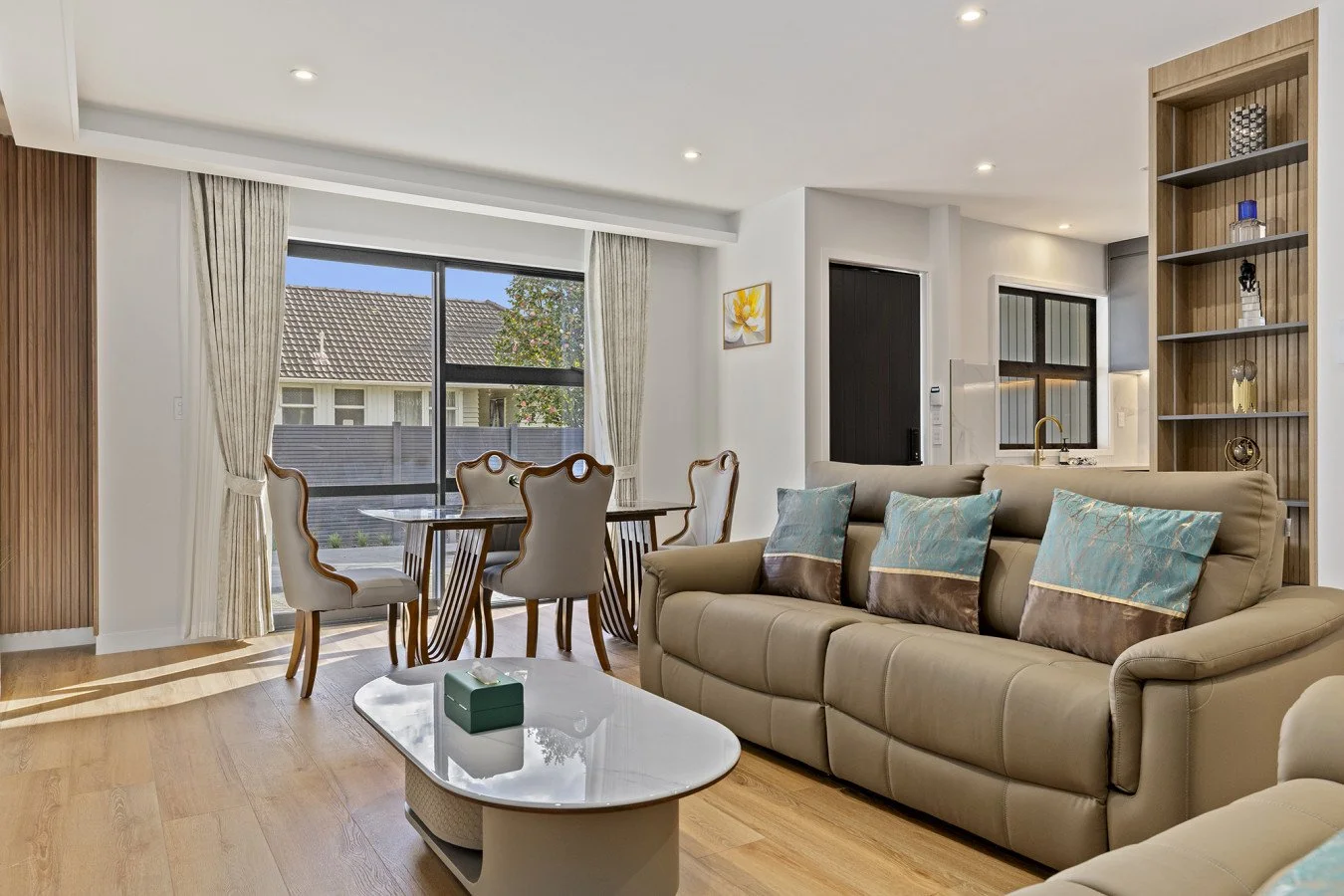 Living room with a beige sofa, blue and brown pillows, a white coffee table, wooden dining table with six chairs, large window with curtains, view of neighboring houses, built-in wooden shelves, and a small kitchen area.