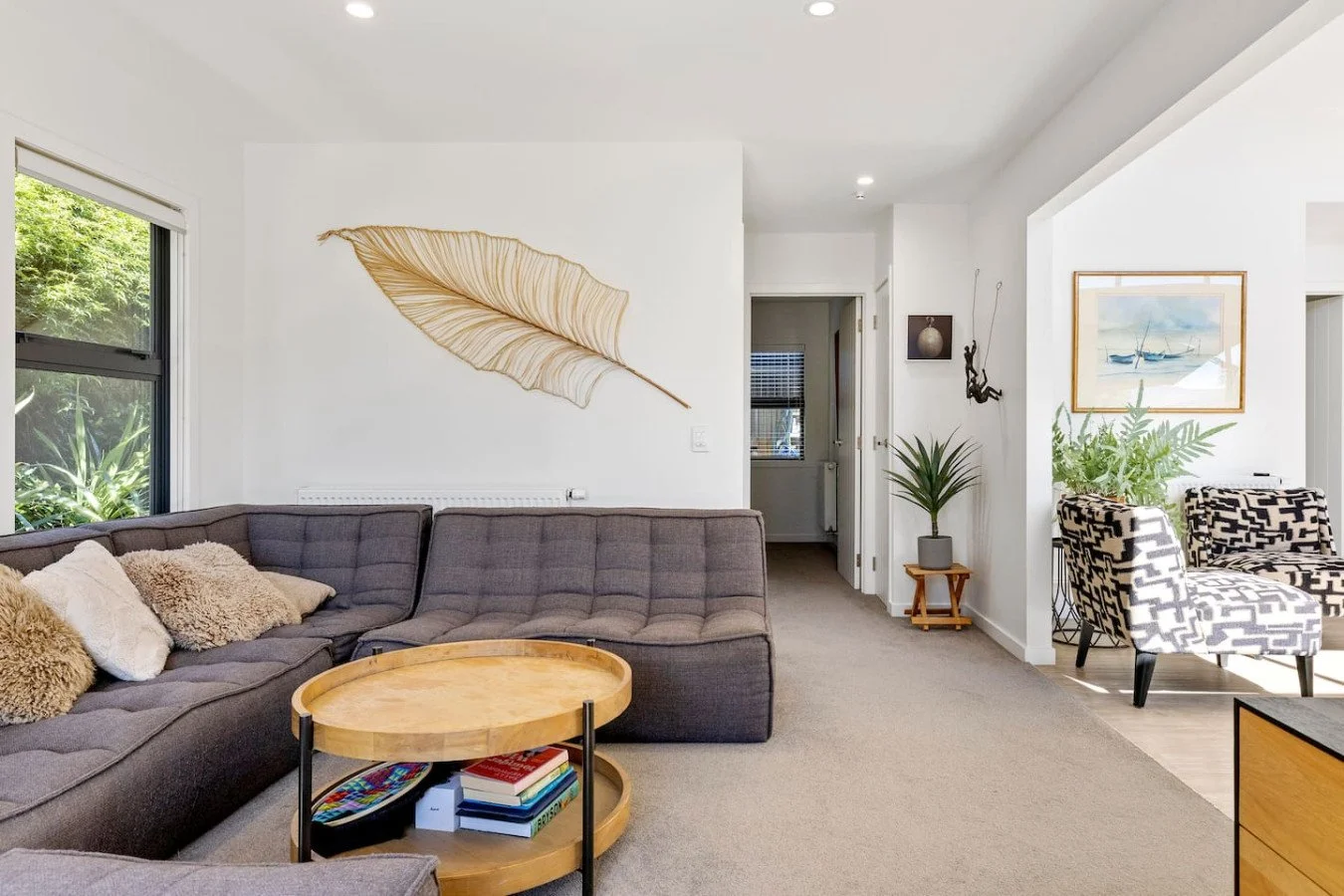 Living room with gray sectional sofa, round wooden coffee table, and patterned armchairs. Wall art includes a large leaf and picture of sailboats. Indoor plants and natural light from windows.